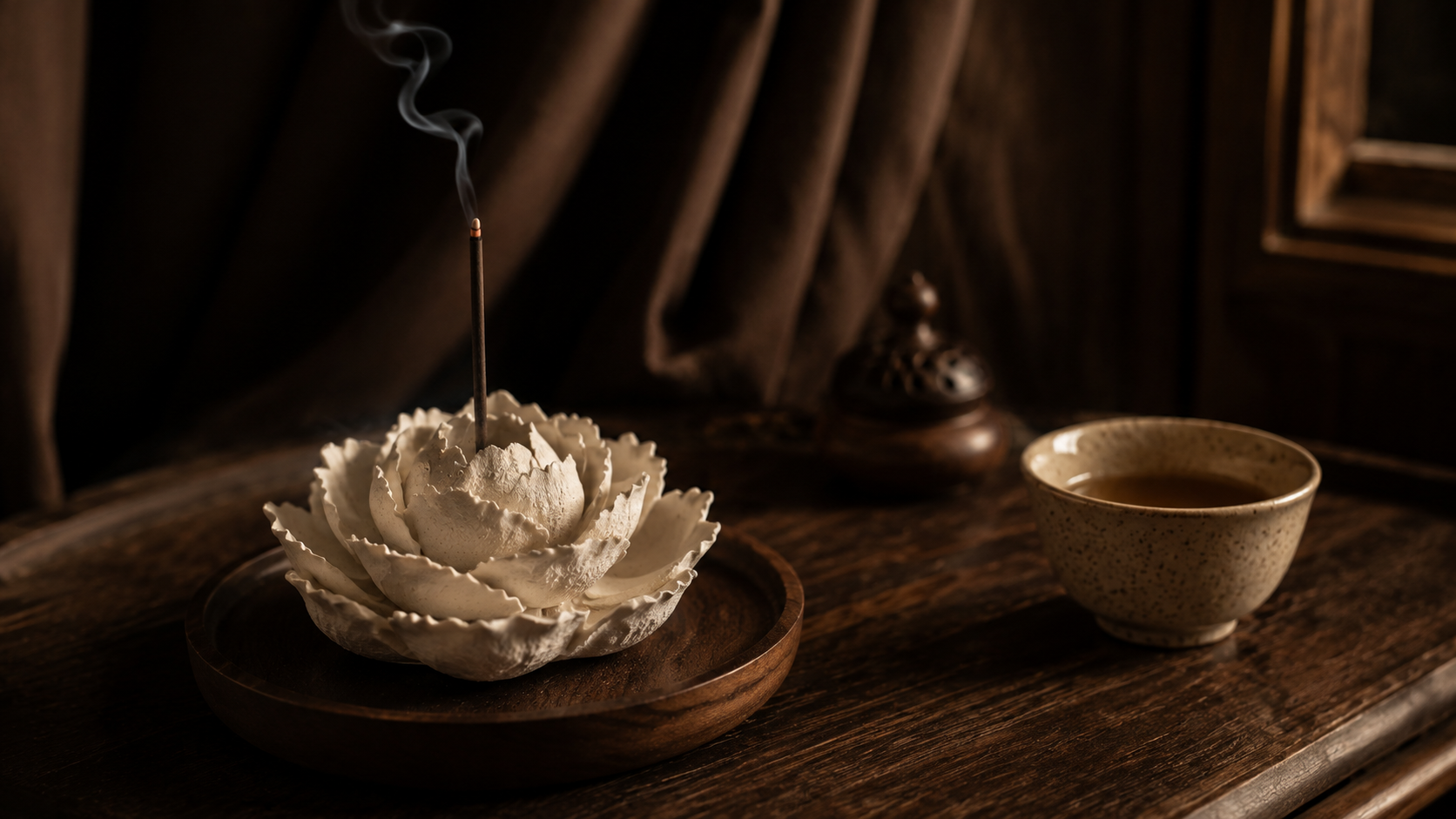 Handcrafted porcelain flower incense burner with rising smoke beside a tea cup on a dark wooden table