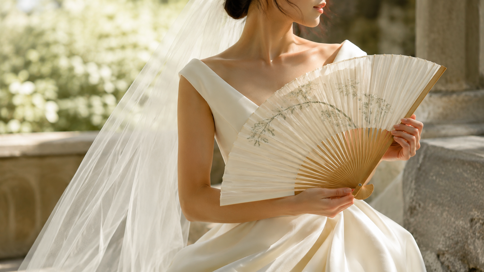 Bride holding a handcrafted silk folding fan beside an ivory wedding dress in soft natural light